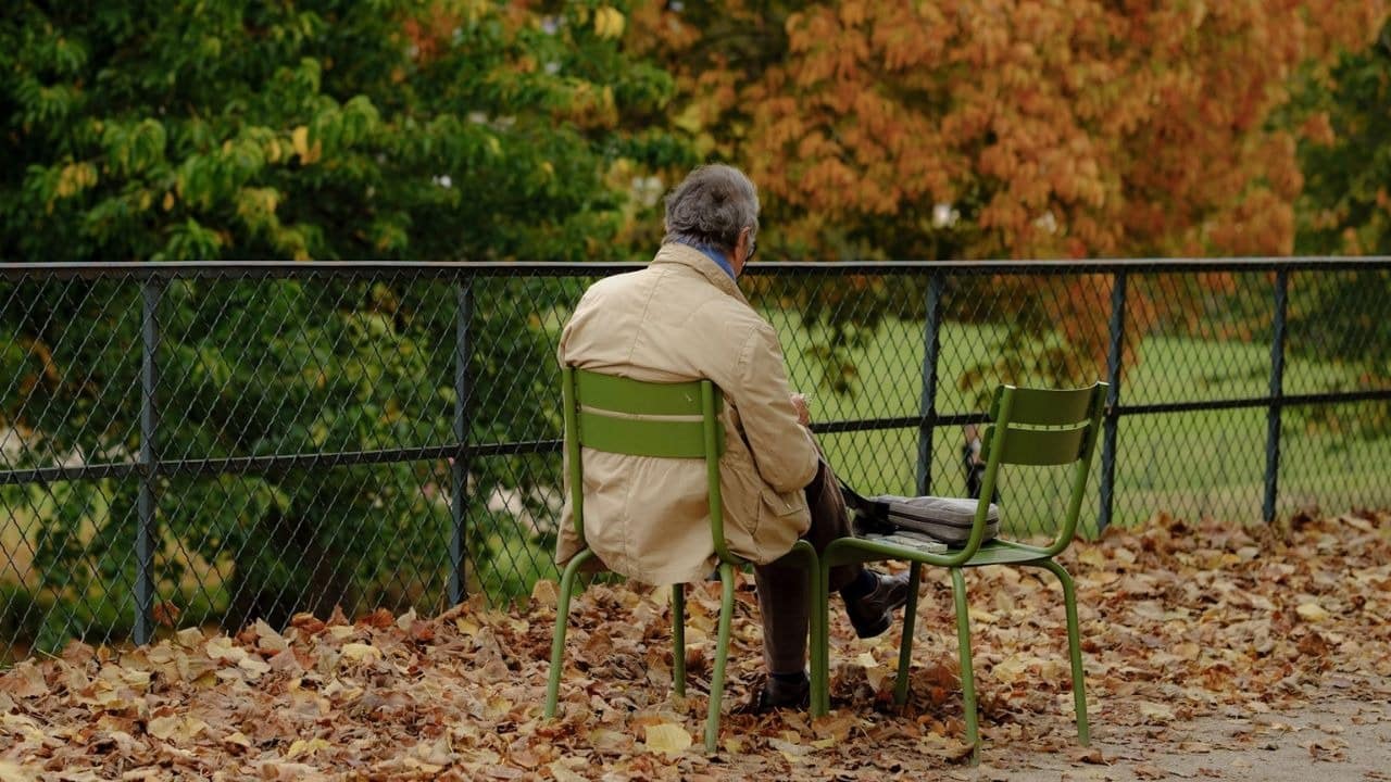 A man in a tan jacket sits alone on a green chair amidst fallen leaves.
