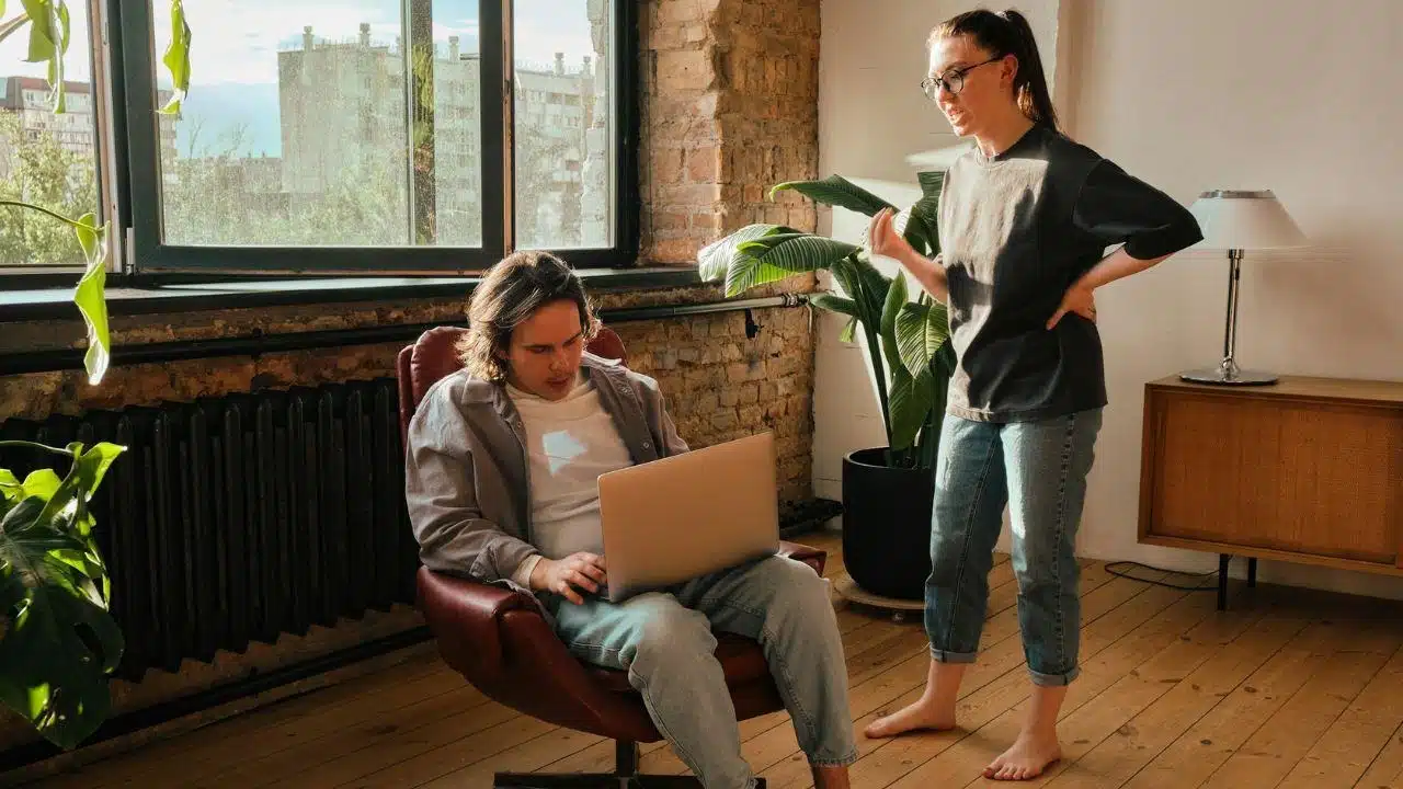 A woman with hands on her hips confronts a man sitting in a chair.