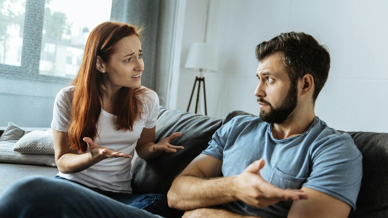 A woman arguing with a bearded man who gestures with his hand as they sit on a couch.