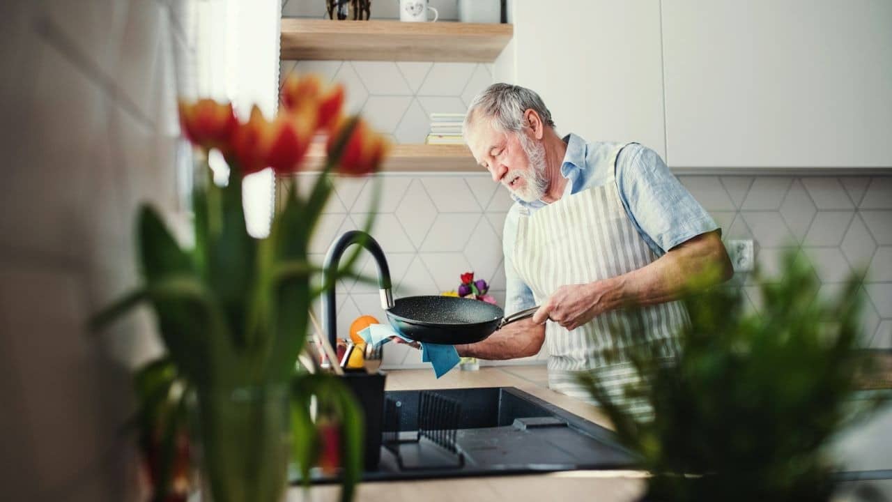 A bearded man in an apron washes a frying pan at a kitchen sink.