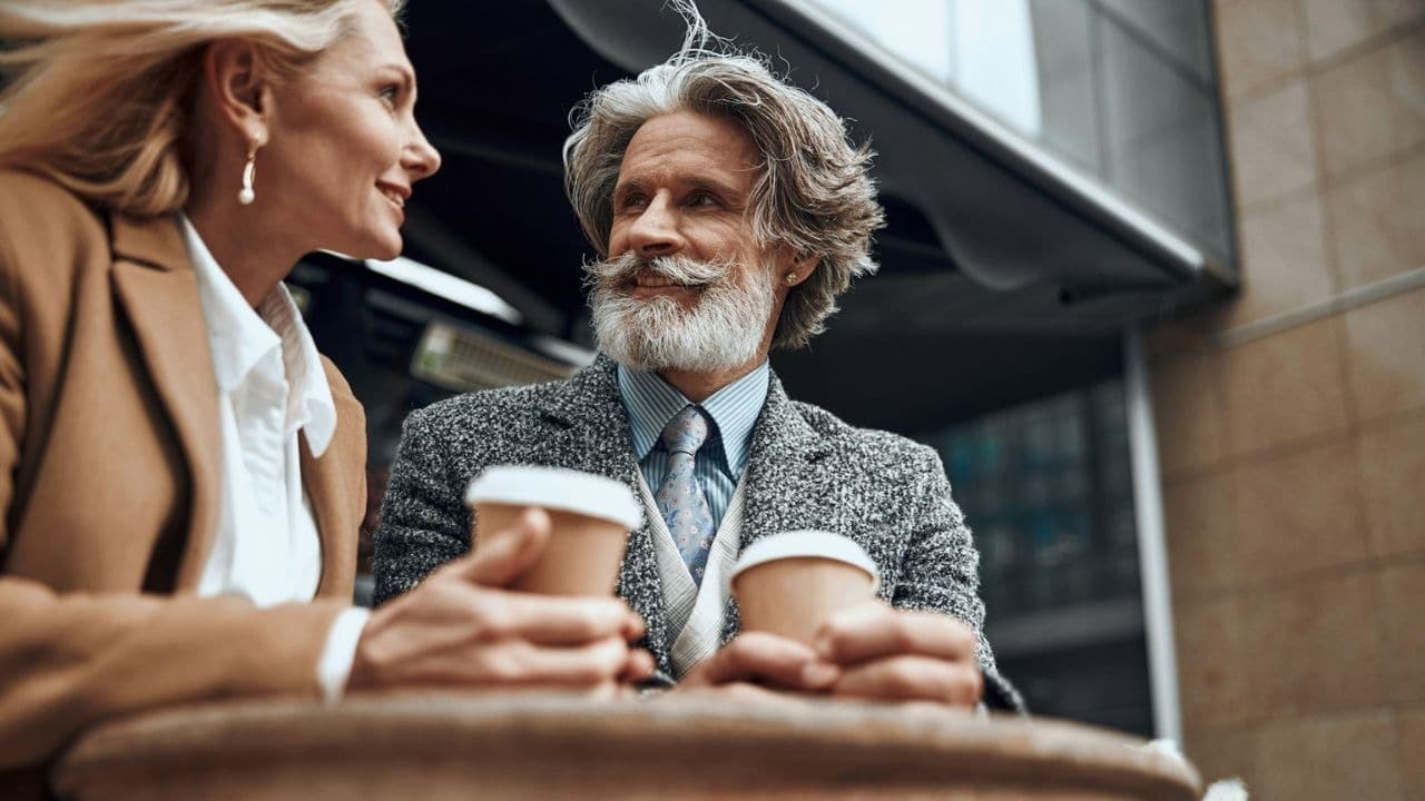 A stylish older couple sits outdoors, each holding a coffee cup.