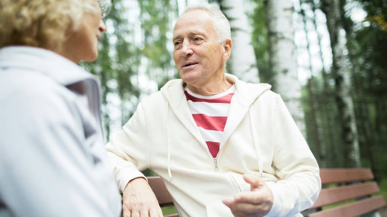A man in a striped shirt and white hoodie talks to a woman on a bench.