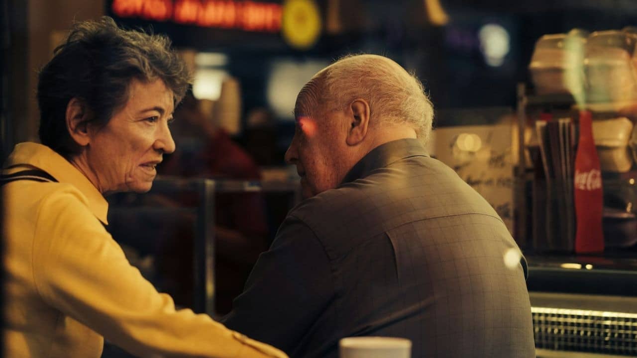 A woman in a yellow shirt talks to a man with white hair in a dimly lit diner.