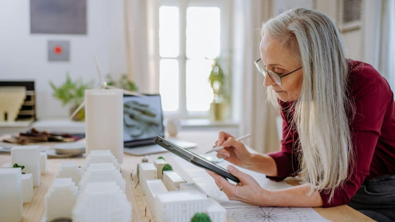 A gray-haired woman in glasses works on a tablet at a desk.
