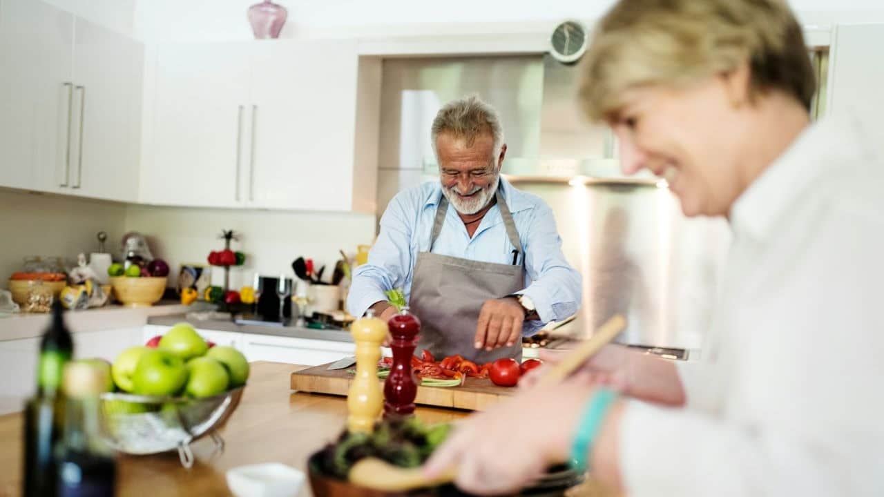 A man in an apron chops vegetables in a kitchen while a woman in the foreground prepares food.