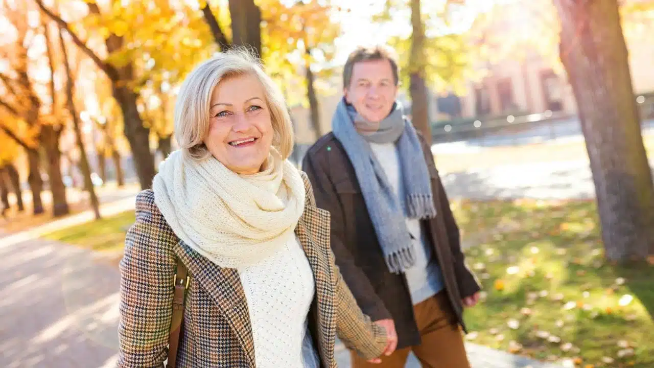 A smiling older couple holds hands while walking outdoors in autumn.