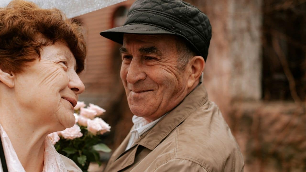 A senior man in a cap smiles at a woman holding flowers.