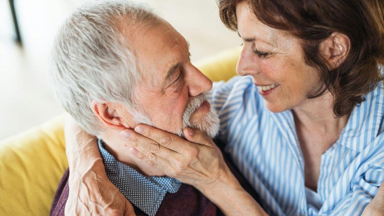 A smiling woman with brown hair embraces a bearded man, looking into his eyes.