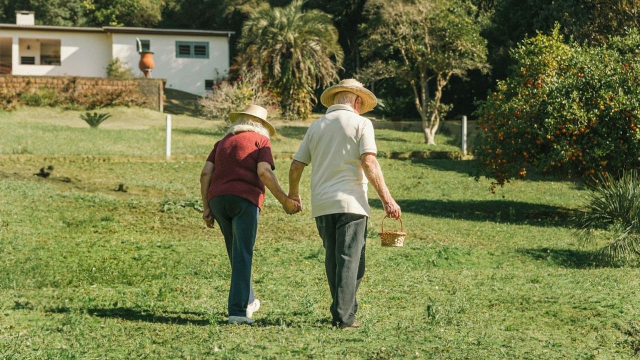 A senior couple wearing straw hats walks hand-in-hand across a grassy field.