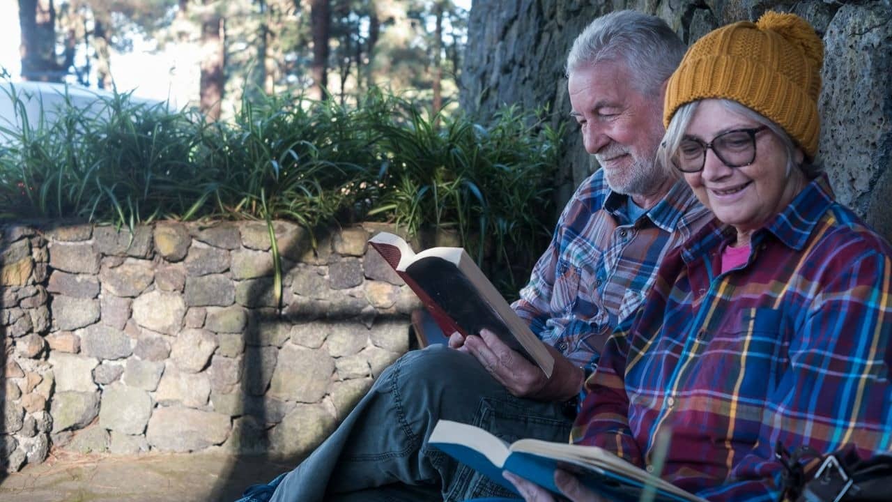 A senior couple sits outdoors, reading books and smiling.