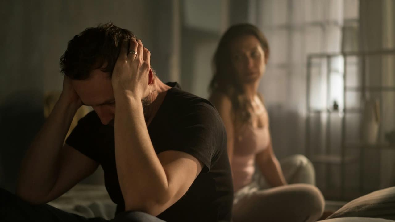 A man sits in the foreground with his head in his hands, looking distressed, while a woman sits blurred in the background on the bed.