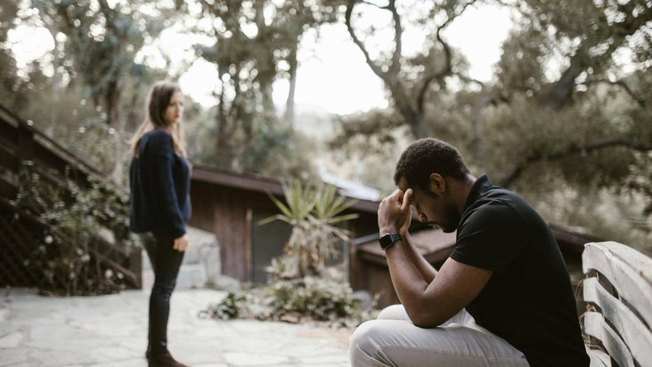 A man sitting with his head in his hands while a woman stands nearby looking upset.