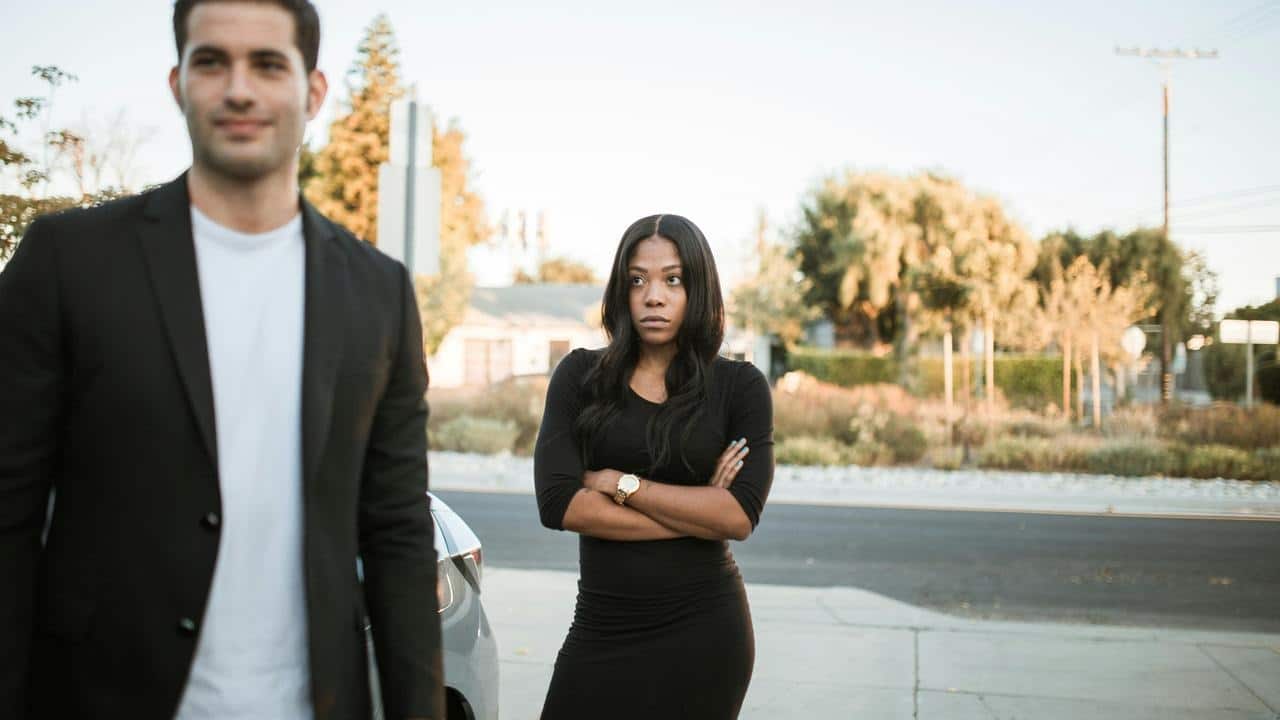 A woman standing with her arms crossed while a man walks away outdoors.
