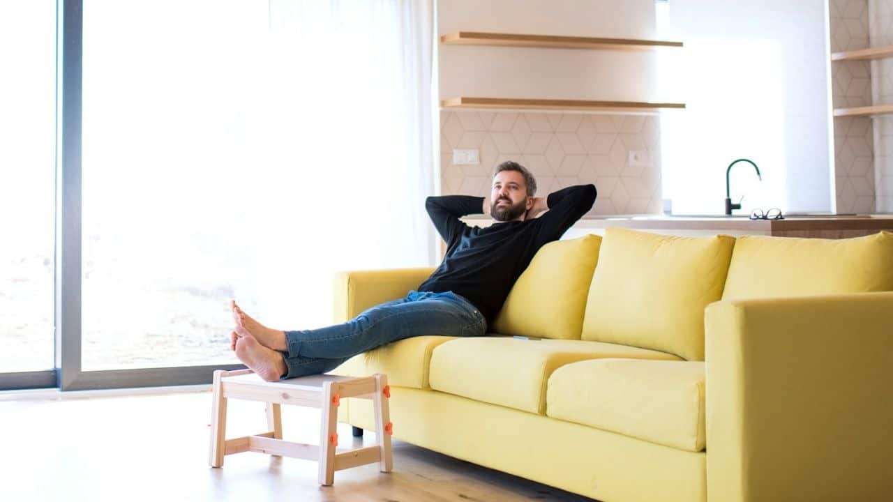 A calm man with a beard is relaxing on a yellow couch with his feet propped up on a stool.