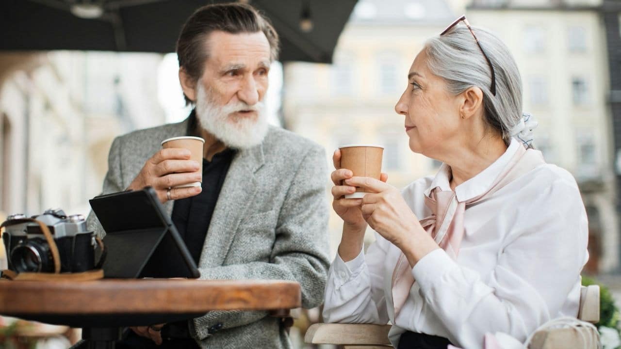 Older man and woman sitting outdoors with coffee cups and a tablet on a table.