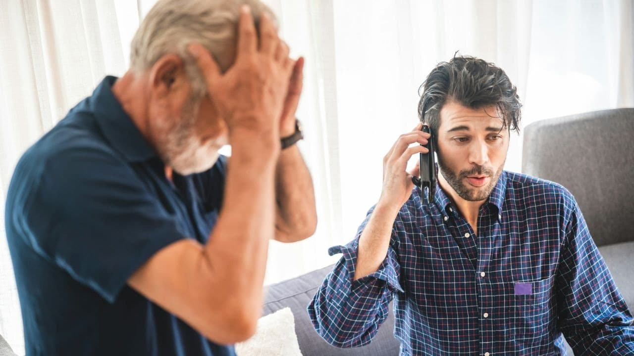 A younger man with a beard is on a phone call while an older, out-of-focus man holds his head in the background.