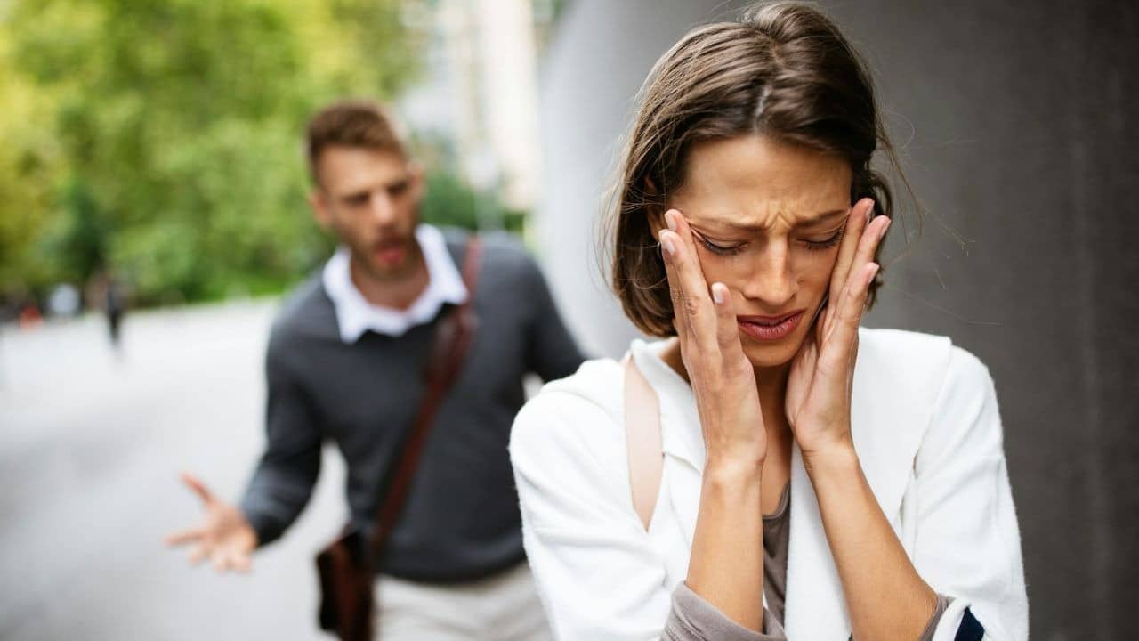 Woman holds her temples in distress while a blurry man shouts and gestures in the background.