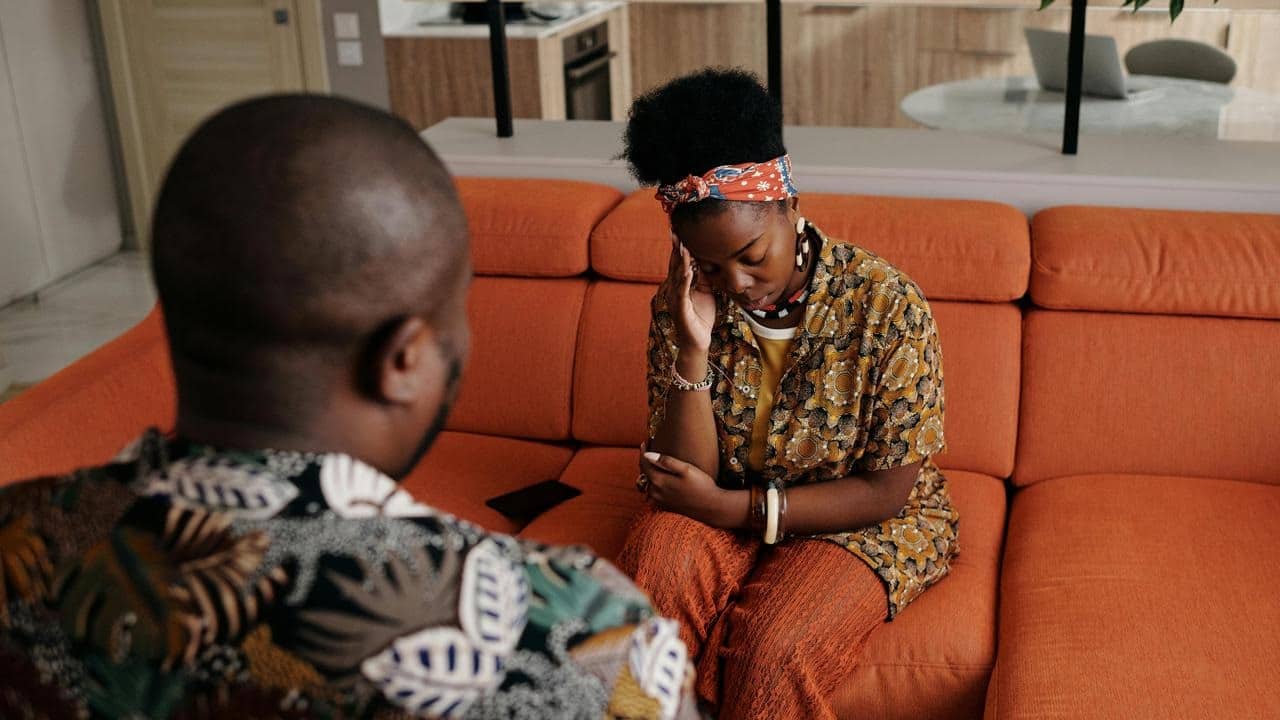 A woman sitting on an orange couch looking upset while a man sits across from her.