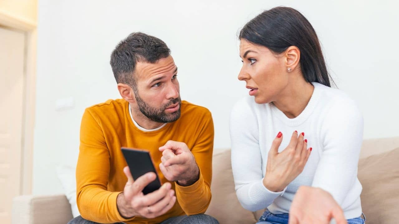 Man holds a phone and speaks to a woman who is gesturing defensively on a sofa.