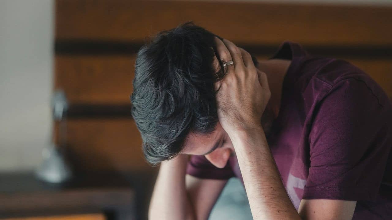 A man with dark hair sits with his hands on his head, looking distressed.