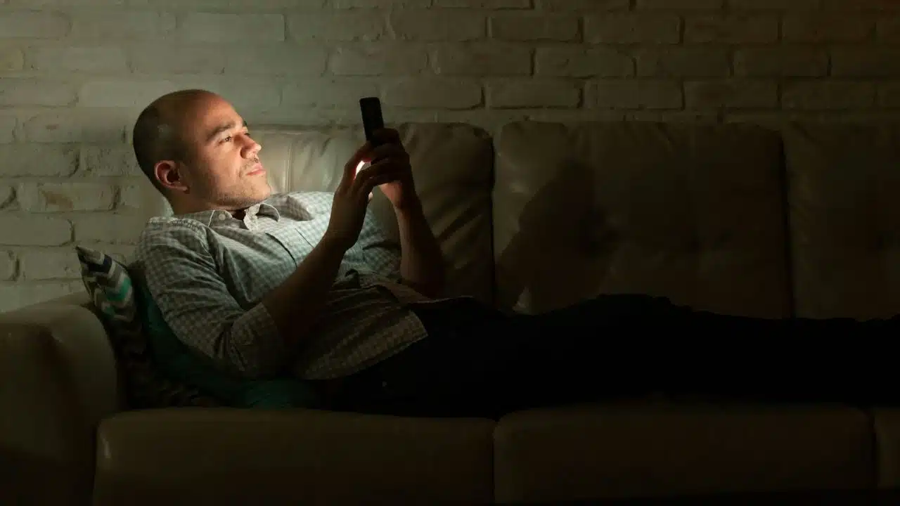 A bald man lies on a couch in a dark room, intently looking at his phone.