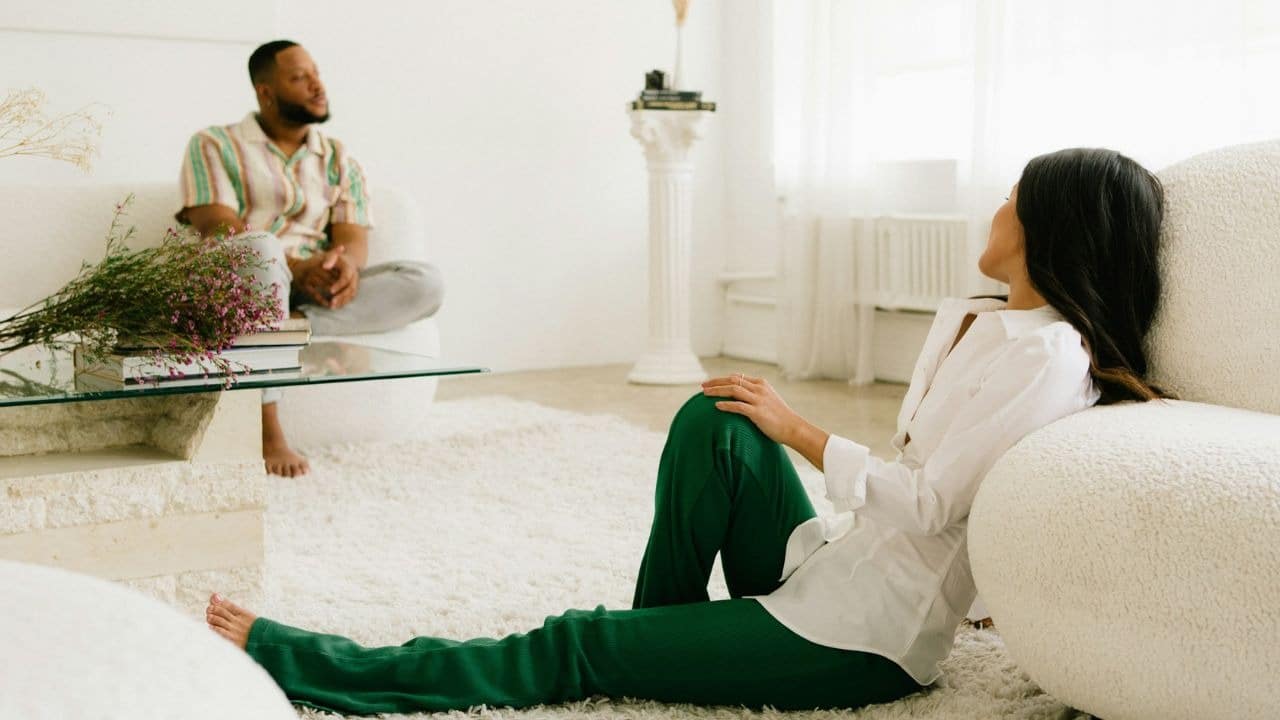 A woman in green pants and a white shirt leans against a sofa, while a man sits on another sofa in the background.