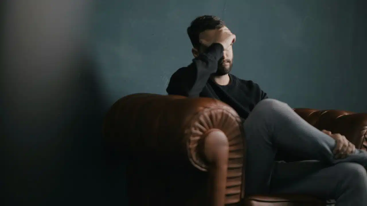 A distressed man with a beard sits on a brown leather couch, hand covering his face.