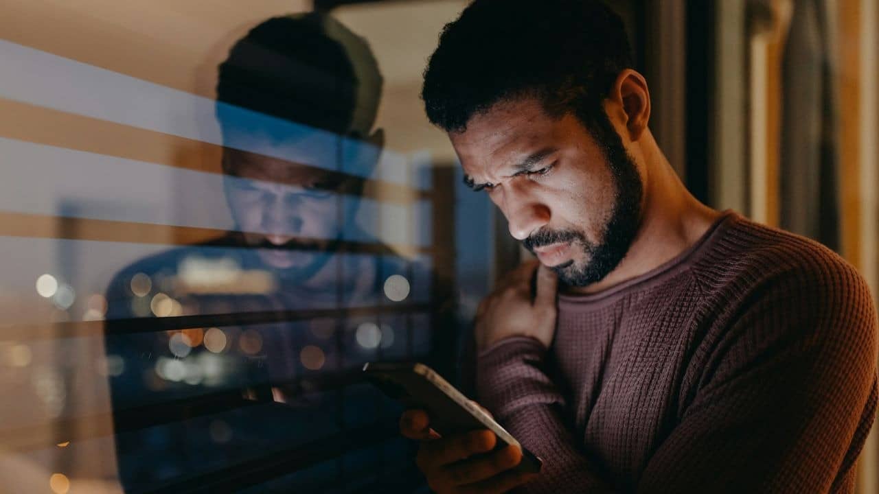 A concerned man looks at his phone while his reflection is visible in the window.