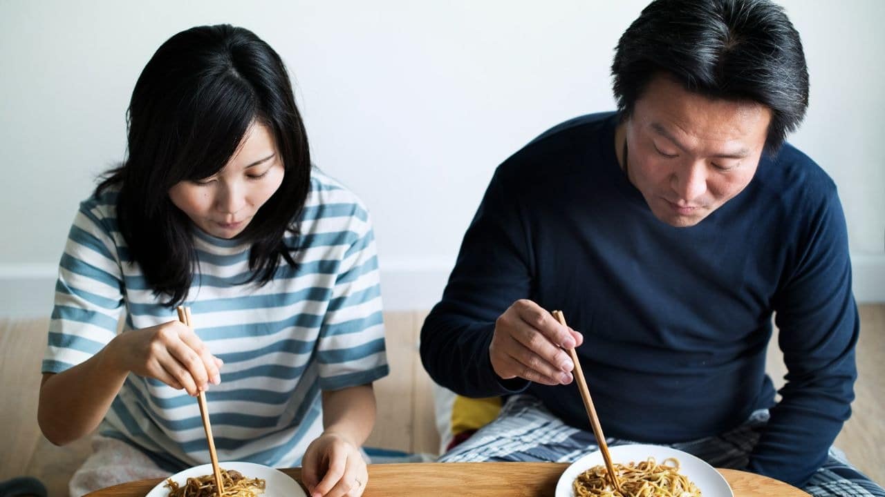 A man and a woman sit on the floor, eating noodles with chopsticks.