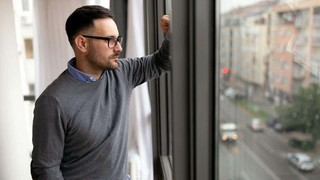 A man wearing glasses and a grey sweater looks out a window thoughtfully.