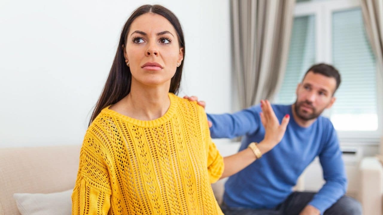 A woman in a yellow sweater looks away with a distressed expression as a man reaches out.