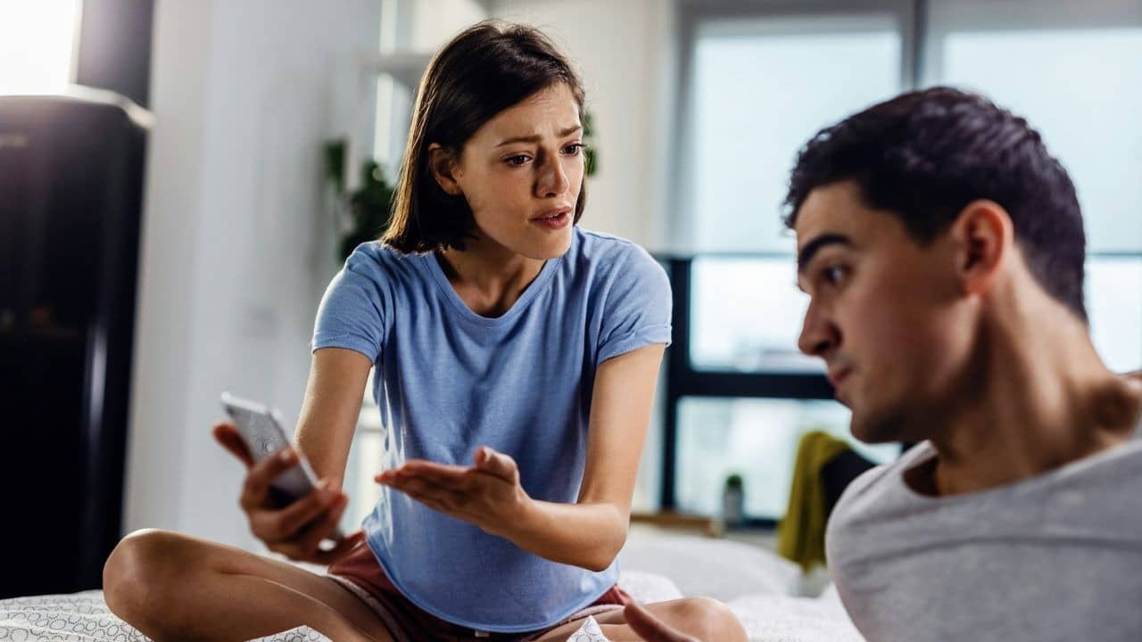 A distressed woman holding a phone gestures towards a man, seemingly arguing.