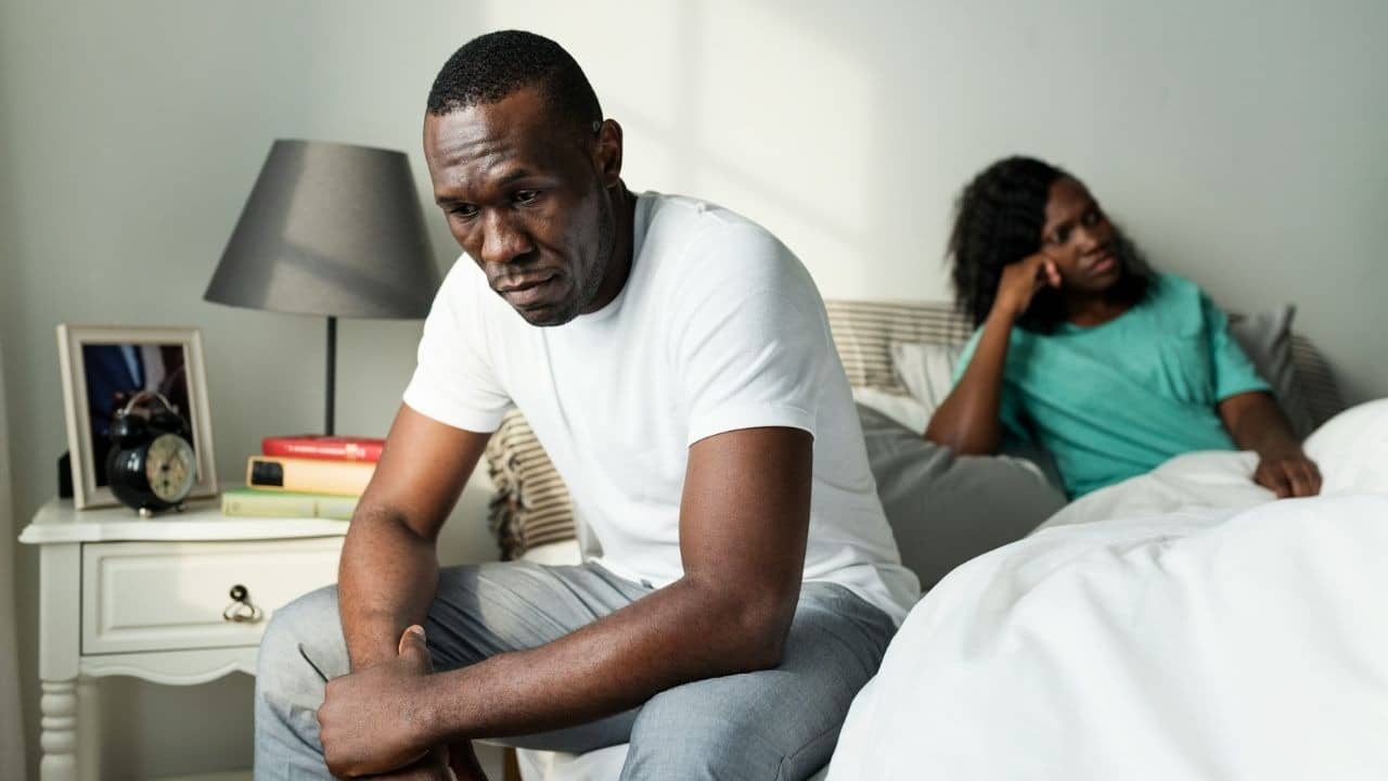 A dejected man sits on the edge of a bed, while a woman looks away in the background.