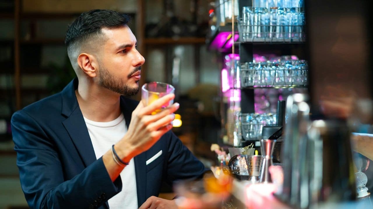 A man with a beard in a dark jacket sips a drink at a bar.