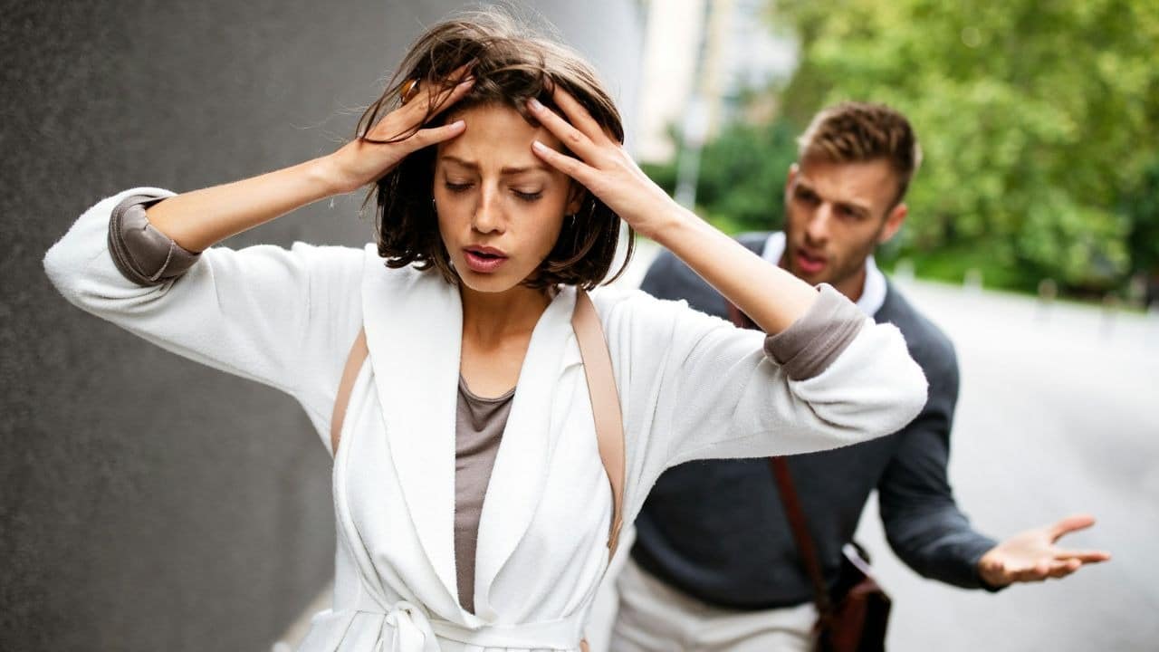 A distraught woman holds her head while a man gestures angrily behind her.