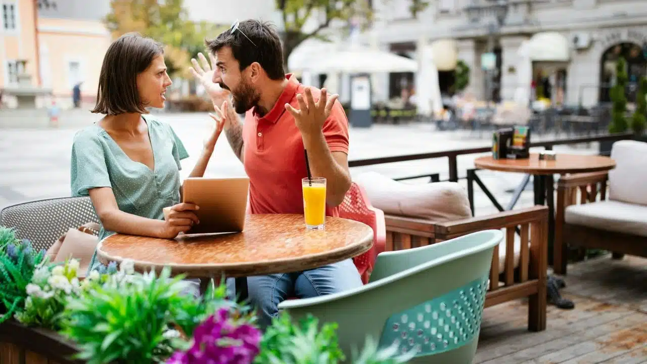 A man dramatically gestures while arguing with a woman holding a tablet at a cafe.