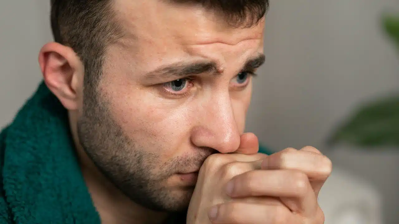 A close-up of a man with blue eyes and a scruffy beard, his hands clasped under his chin.