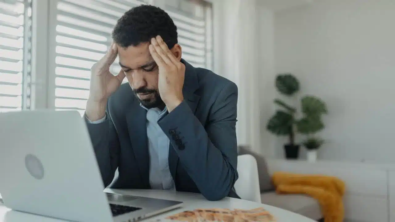 A distressed man with his hands on his head sits at a table with a laptop and money