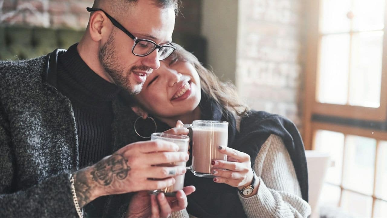A smiling couple in a cafe clinks clear mugs of a light brown beverage.