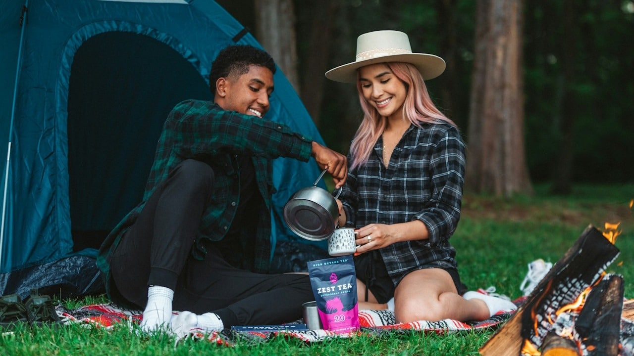 A man pouring tea in the cup of his girlfriend while they are sitting in the grass during camping.