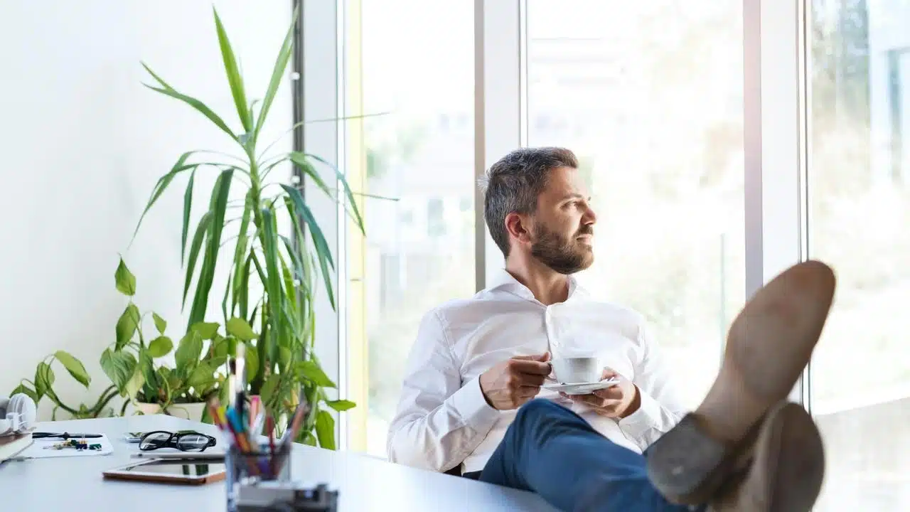 A bearded man in a white shirt with his feet on a desk holding a coffee cup.