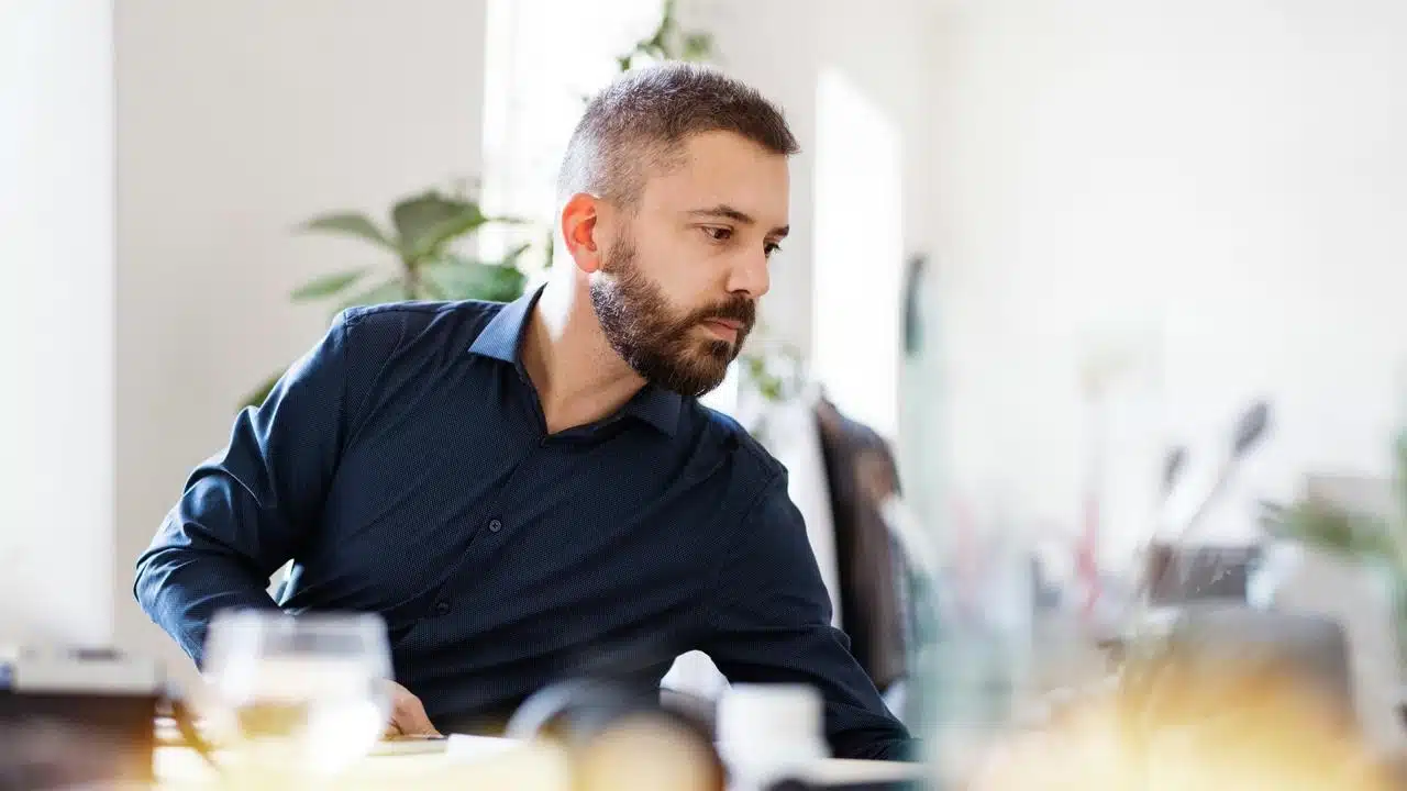 A bearded man in a dark shirt reaching across a desk.