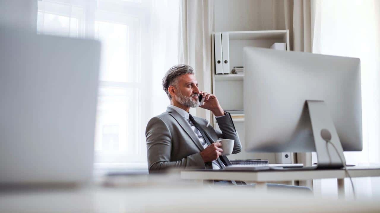 A man in a suit at his desk on a phone call holding a mug.