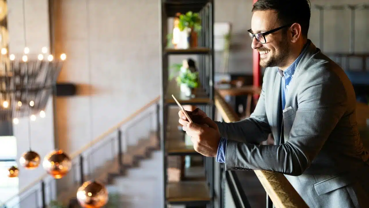 A smiling man in a suit leaning on a railing while using a smartphone.