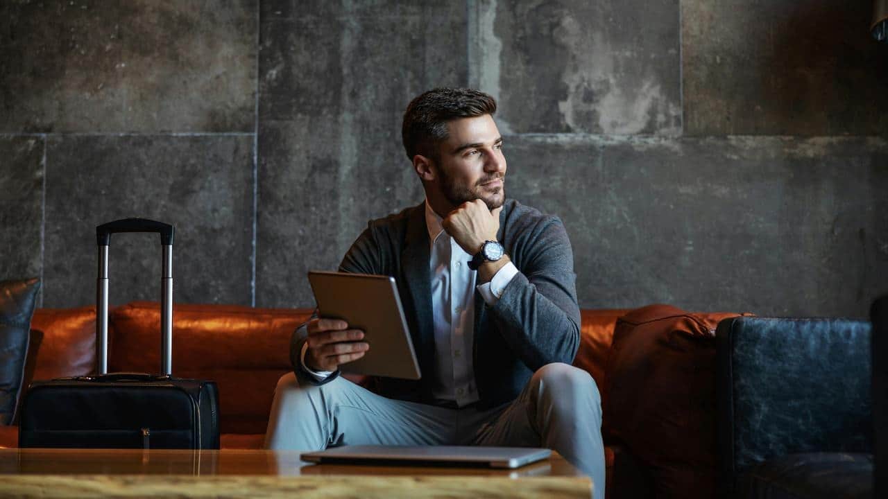 A businessman sitting on a couch holding a tablet with a rolling suitcase beside him.