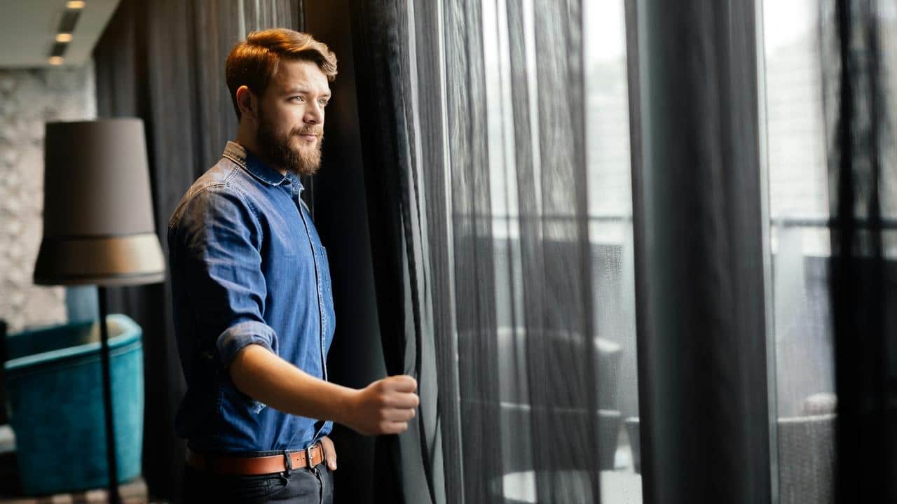 A bearded man in a denim shirt pulling back curtains.
