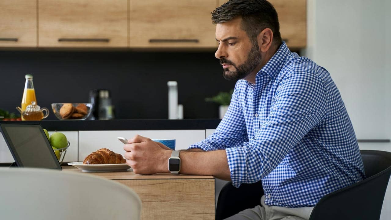 A man in a checkered shirt at a kitchen table using his phone.