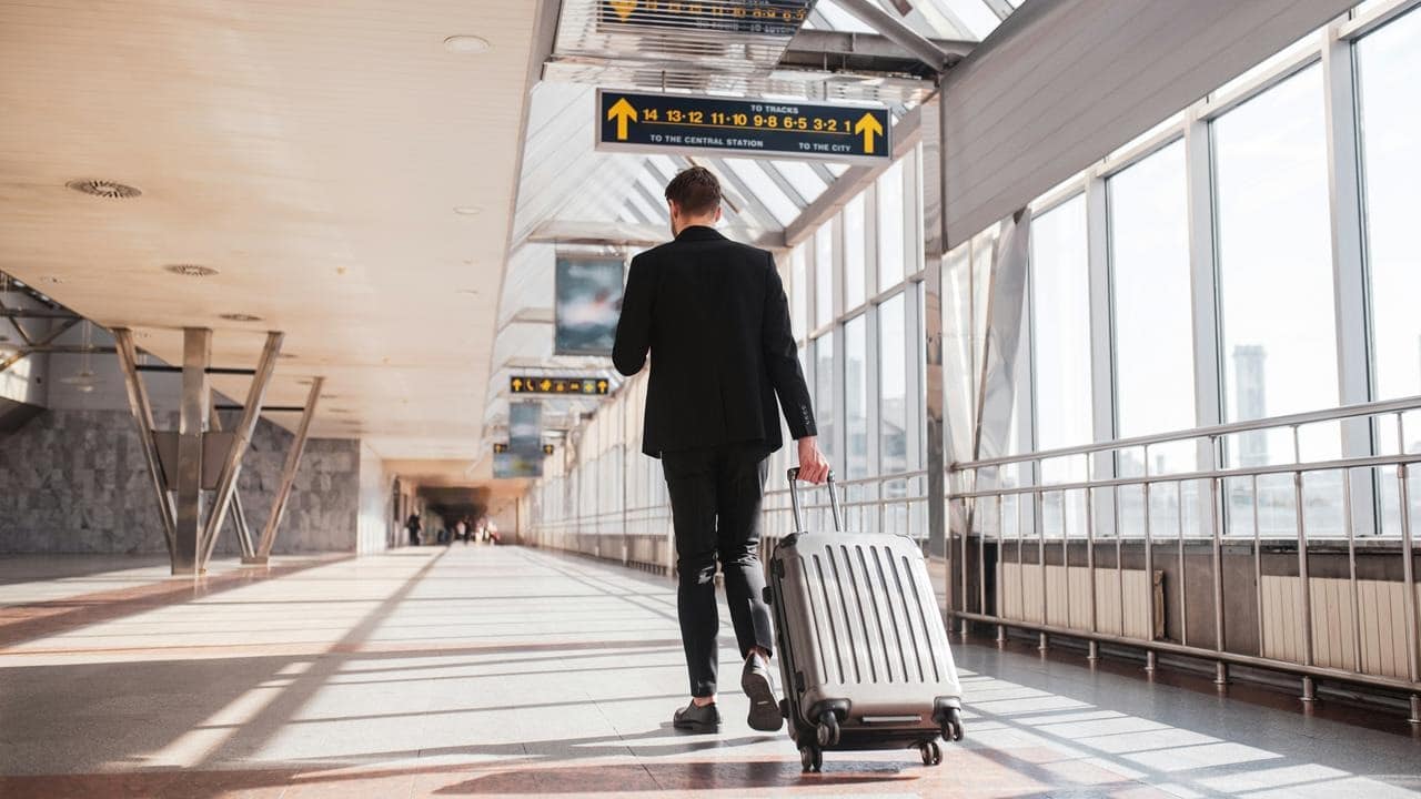 A suited man walking through a station terminal pulling a rolling suitcase.