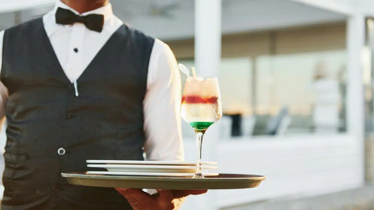 A waiter holding a tray with stacked plates and a layered cocktail.
