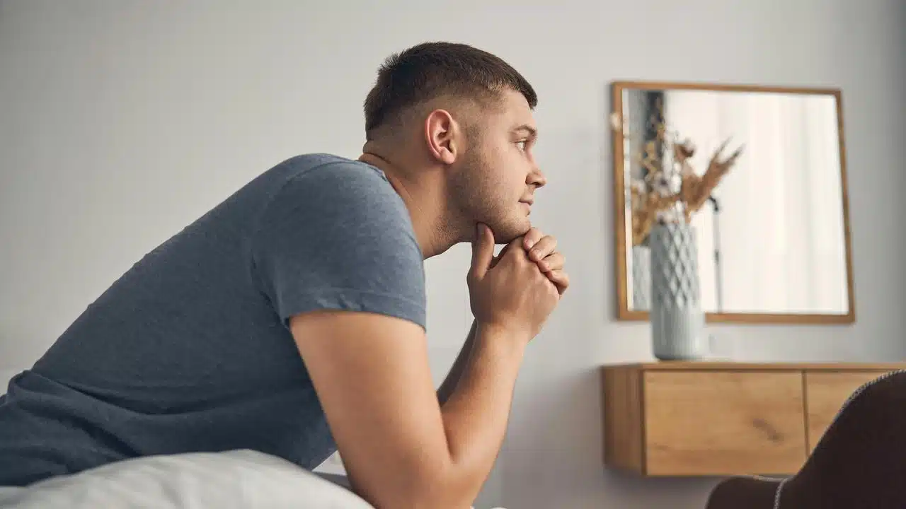 A man sitting on a bed leaning forward with hands clasped under his chin.